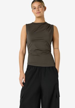 Jeune femme portant un haut kaki foncé sans manches et un pantalon cargo noir ample, debout devant un fond blanc.