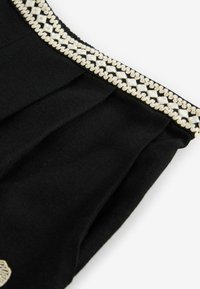 Tissu noir avec des plis, présentant une bordure brodée blanche et beige avec des motifs géométriques le long du bord.