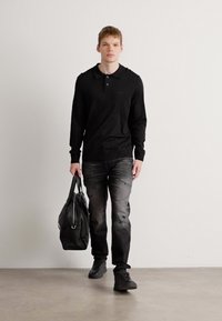 Cămașă polo neagră cu mânecă lungă, blugi închiși la culoare cu o ușoară decolorare, teniși negri și o geantă de voiaj din piele neagră. Fundal alb simplu.
