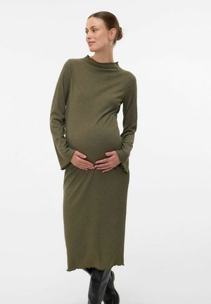 Robe de maternité vert olive à manches longues, texture côtelée et ourlet festonné. Le modèle berce son ventre de femme enceinte, portant des bottes noires.