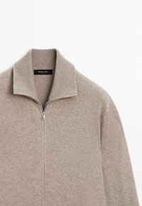 Ljus taupe zip-up tröja med hög krage, tillverkad av mjukt tyg, med ribbade detaljer vid kragen och dragkedjan. Etikett synlig inuti.