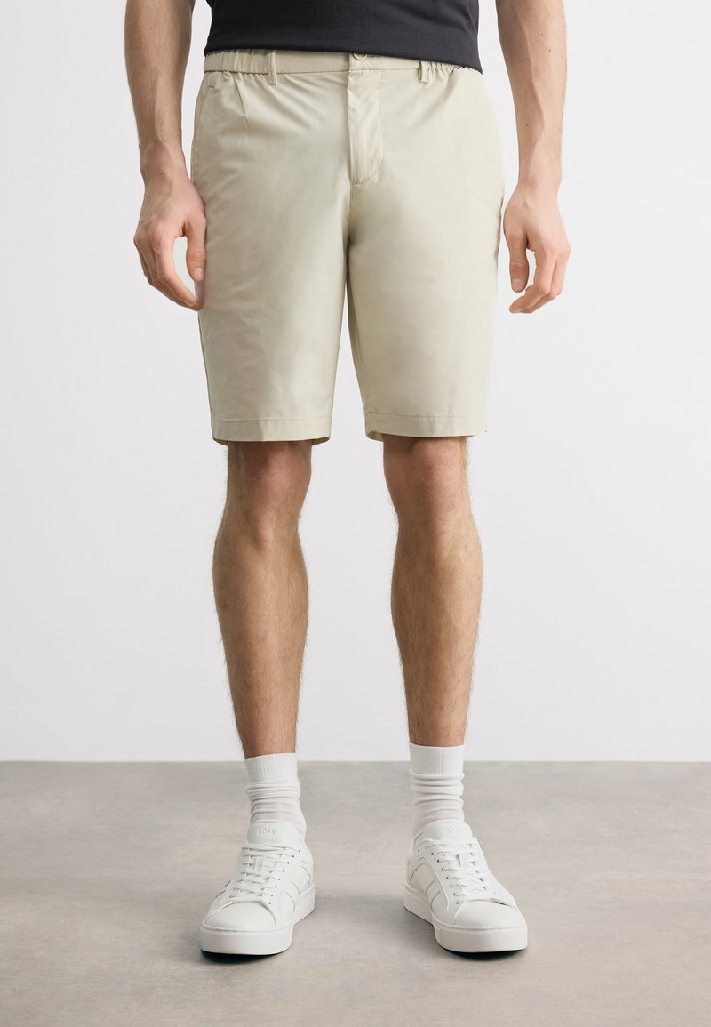 Άνδρας που φοράει μπεζ σορτς μέχρι τα γόνατα, λευκές κάλτσες μέχρι τον αστράγαλο και λευκά αθλητικά παπούτσια, στέκεται μπροστά σε απλό ανοιχτό φόντο.