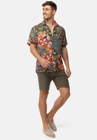 Camisa de manga corta de algodón con un colorido patrón geométrico en verde, naranja y púrpura. Combinada con pantalones cortos verde oliva y zapatos cómodos grises.