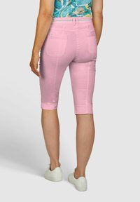 Helles rosa Capri-Hosen mit zwei Gesäßtaschen, einem taillierten Schnitt und einem umgeschlagenen Saum. Das Modell trägt weiße Sneaker für einen lässigen Look.