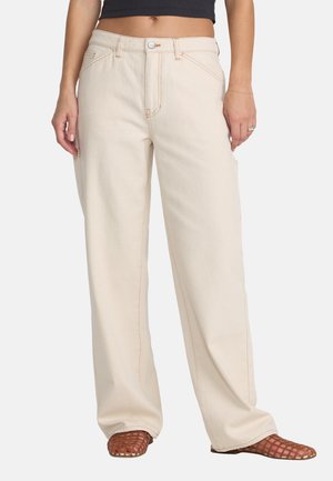 Vrouw die beige broek met wijde pijpen, bruine gevlochten schoenen en een zwart cropped top draagt, staand tegen een effen achtergrond.