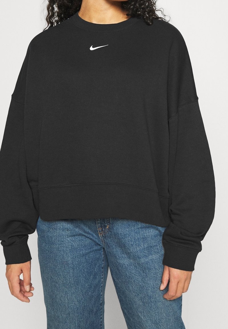 Black sweatshirt with a crew neckline and oversized fit. Features a white Nike logo on the chest. Paired with blue jeans.