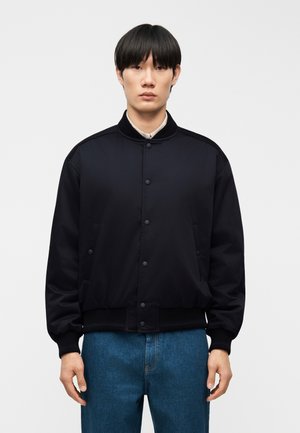 Jeune personne aux cheveux noirs et lisses portant une veste noire boutonnée et un jean bleu, se tenant devant un fond clair uni.