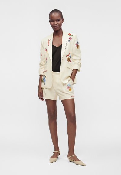 Femme debout et souriante, portant un blazer crème et un short avec broderies colorées, un haut noir et des ballerines beiges à bout pointu.