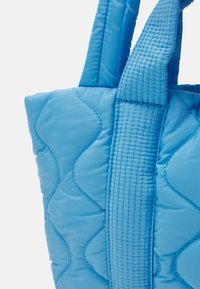 Blauwe gestepte tote bag met golvende stikpatronen, gewatteerde textuur en brede handvatten met lineaire stikdetails.