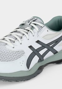Λευκό και γκρι αθλητικό παπούτσι Asics με μαύρες λωρίδες λογότυπου, λευκά κορδόνια και πράσινη μαξιλαρωτή σόλα, γωνιακά τοποθετημένο ώστε να φαίνονται λεπτομέρειες από το πλάι και το μπροστινό μέρος.