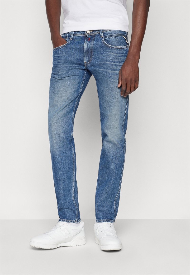 Homme portant un jean bleu slim avec un léger délavage et des baskets blanches, debout avec une main dans la poche devant un fond uni.