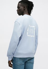 Svetlomodrá mikina s okrúhlym výstrihom a bielou grafickou potlačou na chrbte, vyrobená z mäkkej látky, s rebrovanými manžetami a lemom.