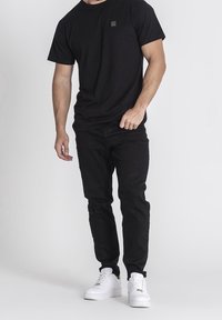 Camiseta de algodón negra con un pequeño logo en el pecho, combinada con jeans negros y zapatillas blancas, presentando un atuendo casual.