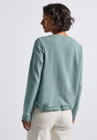 Femme aux cheveux courts et bouclés, portant un haut à manches longues teal clair et un pantalon blanc cassé, de dos contre un fond clair.