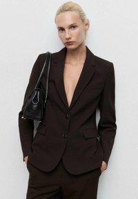 Hnědý na míru střižený blazer s hlubokým výstřihem do V, jednoduchým knoflíkovým zapínáním a dvěma předními kapsami. Nosit s černou kabelkou přes rameno.