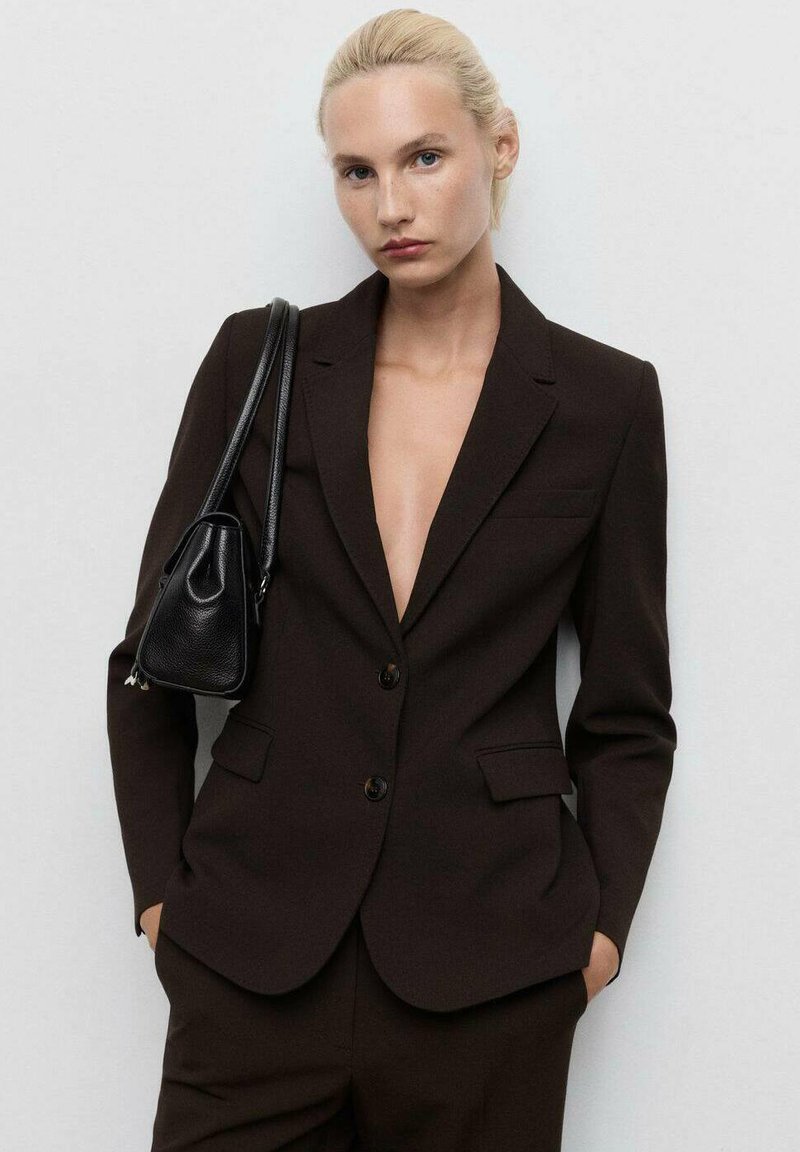 Hnědý na míru střižený blazer s hlubokým výstřihem do V, jednoduchým knoflíkovým zapínáním a dvěma předními kapsami. Nosit s černou kabelkou přes rameno.