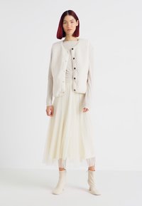 Hvitt strikket vest med svarte knapper over en lys beige topp og en plisserte, ankel-lange, kremhvite skjørt; kombinert med beige hæler.