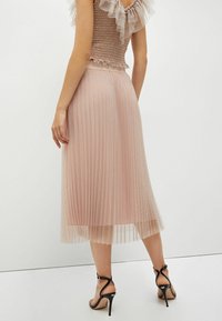 Mujer con una falda midi plisada de color rosa empolvado y una blusa con volantes, combinadas con sandalias de tacón alto negras, de pie frente a una pared blanca.