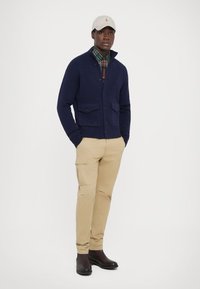 Mornarska cardigan s gumbnimi zaprtji in dvema žepe, ležerna preko karirane srajce. Bež cargo hlače in temni škornji dopolnjujejo izgled.