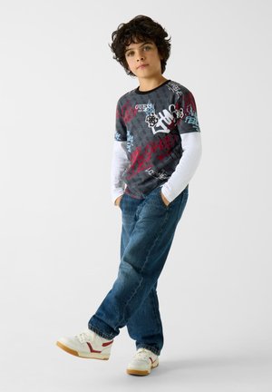 Kurzärmliges Grafik-T-Shirt in Grau mit roten und blauen Designs, über einem weißen Langarmshirt getragen. Kombiniert mit blauen Jeans und weißen Sneakern.