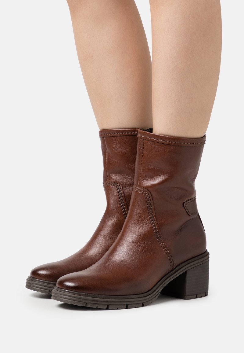 Bottes en cuir marron mi-mollet à talons blocs portées sur jambes nues sur un fond blanc uni.