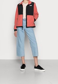 Osoba ubrana w koralową i czarną polarową kurtkę North Face, jasnoniebieskie, krótko przycięte, szerokie dżinsy, białą koszulę i czarne sneakersy z białymi podeszwami.