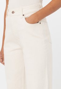 Pantalones anchos de color crema en mezcla de algodón, con cintura alta, bolsillos laterales y detalles de botones metálicos. Tejido suave y texturizado.