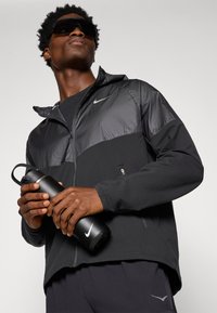 Czarna kurtka Nike z zamkiem z przodu, teksturowana szara wstawka i kaptur. Czarna butelka w ręku. Ciemna koszulka i dresy, nowoczesny sportowy krój.