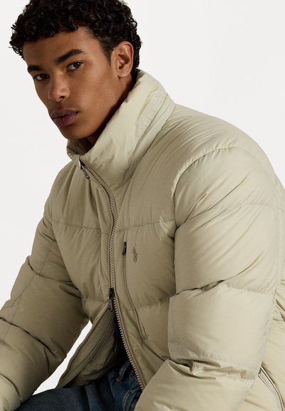 Light beige puffer jacket with a high collar, zip closure, and subtle logo on the chest. Features a quilted texture and padded design.