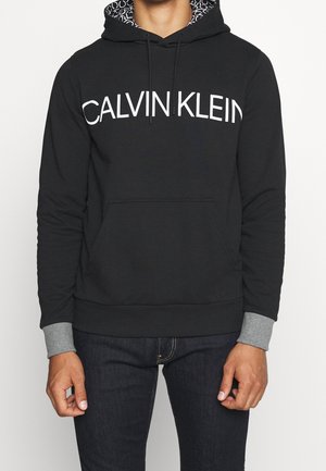 Mann trägt schwarzen Hoodie mit weißem "CALVIN KLEIN"-Schriftzug auf der Brust, grauen Bündchen und gemustertem Kapuzenfutter, kombiniert mit dunkler Jeans.