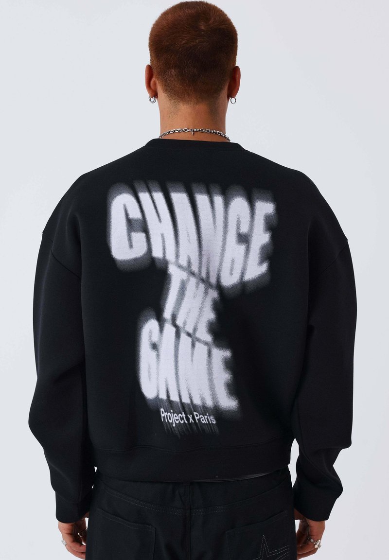 Personne aux cheveux courts portant un sweat-shirt noir avec un texte blanc audacieux et flou "CHANGE THE GAME" et un plus petit "Project x Paris" au dos.