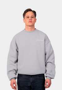 Gai pelēks flīsa krekls ar apaļu kakla izgriezumu un ribotām piedurknēm. Krūšu zonā ir balti uzdrukāts vārds "AIZLIEDZ". Apakšā uzvilktas tumšas džinsas.