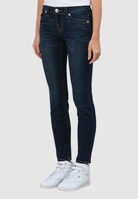 Donkere skinny jeans met een hoge tailleband, voorzien van contrasterende oranje stiksels en zakken aan de voorkant. Gecombineerd met witte hoge sneakers.