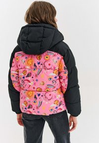 Pufová bunda s ružovým, vzorovaným zadným dielom, na ktorom sú motívy nebeských telies a očí; čierne prešívané rukávy a kapucňa; mäkká, polstrovaná textúra.
