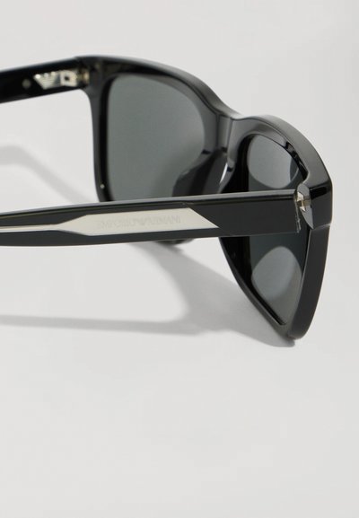 Ochelari de soare din plastic negru cu lentile întunecate, având un design cu cadru geometric și accente argintii pe lateral, etichetați "Emporio Armani."