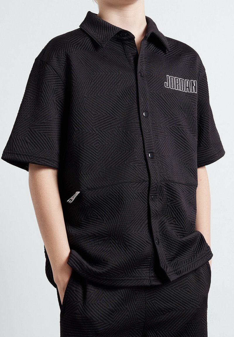Personne portant une chemise noire texturée à manches courtes avec boutons et le texte "JORDAN" sur la poitrine, les mains dans les poches du pantalon assorti.