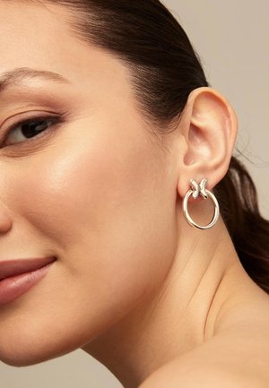 Boucles d'oreilles créoles en argent avec un design accentué torsadé, finition métallique brillante, et forme circulaire, portées à l'oreille d'un mannequin.