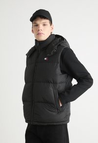 Crna puffer prsluk s sjajnom završnom obradom, s patentnim zatvaračem sprijeda, kapuljačom i logom. Model nosi crnu majicu dugih rukava ispod.