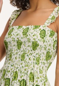 Elastisches Bandkleid mit weißem Grund und grünen Blumenmustern. Rüschenträger und ein figurbetonter Oberteil, der in einen gerafften Rock übergeht.