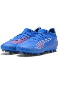 Scarpette da calcio blu con una tomaia testurizzata, caratterizzate da accenti arancioni e un design a strati. Punta arrotondata e tacchetti neri per la trazione.