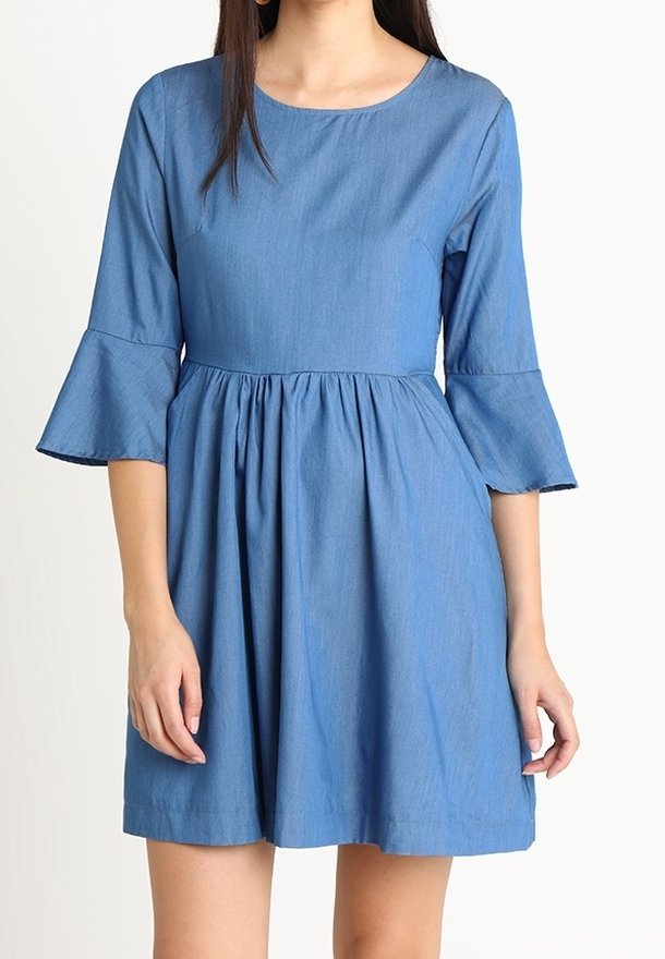 Femme portant une robe bleue mi-longue avec un col rond et des manches évasées trois-quarts, mains reposant sur les côtés.