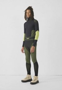 Zwart en neon groen surfpak met een hoge hals, nauwsluitende lange mouwen en gestreepte accenten, gecombineerd met beige wandelschoenen.