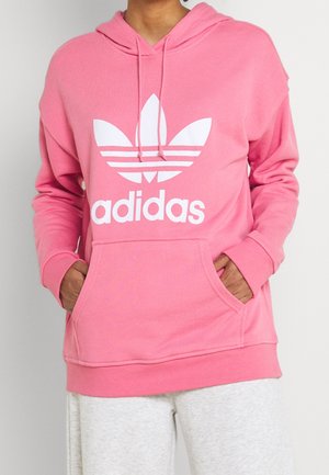 Personne portant un sweat à capuche rose Adidas avec un logo blanc Adidas et les mains dans les poches avant, associé à un pantalon gris clair.