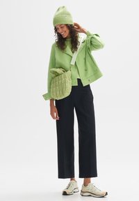 Giacca verde strutturata, berretto abbinato e borsa trapuntata abbinati a pantaloni larghi neri e sneakers chiare con dettagli colorati.