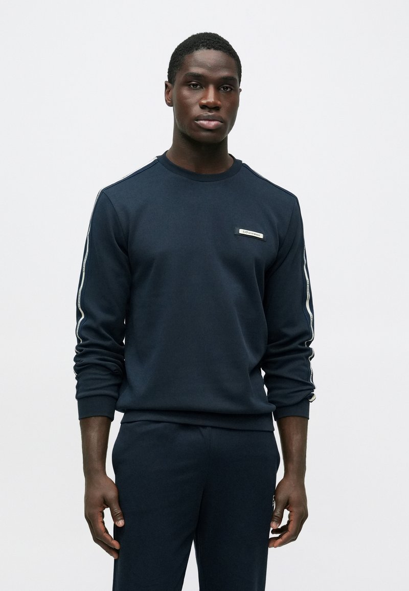 Mladý muž stojaci, oblečený v tmavomodrej mikine a zodpovedajúcich nohaviciach s bielymi pruhmi na rukávoch, na jednoduchom bielom pozadí.