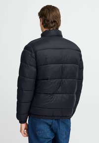 Svart polstret jakke med høy krage, kviltet design og elastiske mansjetter; har en glatt tekstur og løs passform. Bakvinkel vist.