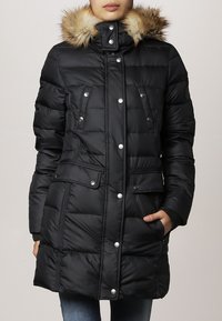 Chaqueta de plumas negra con capucha con borde de piel, botones a presión, bolsillos laterales y acolchado horizontal. Tejido texturizado con puños acanalados.