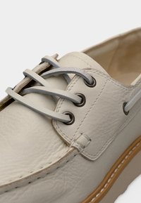 Εγγύς λήψη μπεζ δερμάτινου υποδήματος τύπου μποτάκι με γκρι δερμάτινα κορδόνια διαπερασμένα από μεταλλικούς οπές και καφέ ραμμένη σόλα.