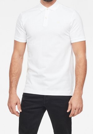 Hombre con una camiseta polo blanca lisa de manga corta con tres botones, combinada con pantalones negros, mostrado desde el cuello hasta la mitad del muslo.