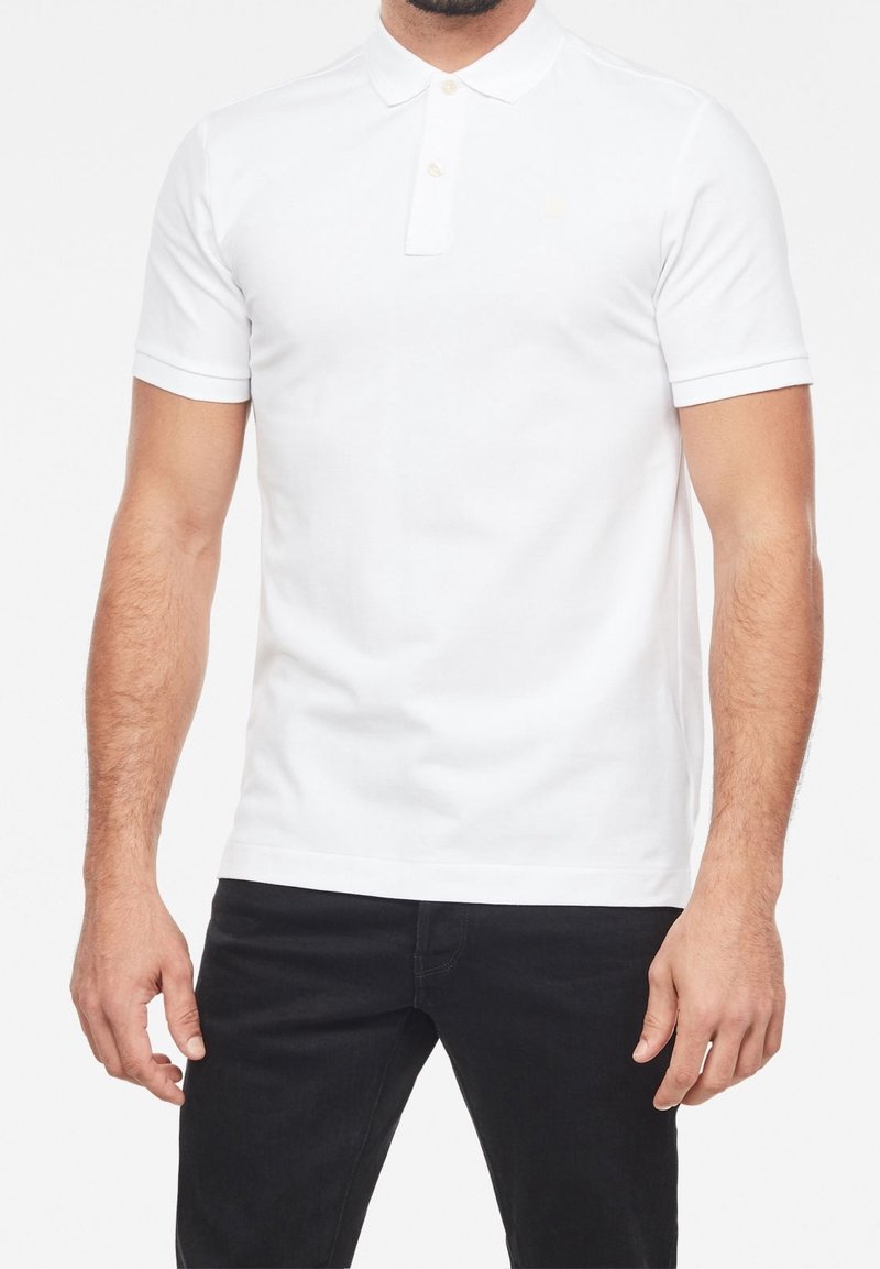 Homme portant un polo blanc uni à manches courtes avec trois boutons, associé à un pantalon noir, visible du cou jusqu'à mi-cuisse.